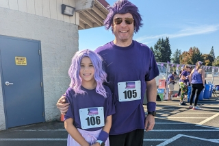 An adult and child dressed in purple wigs and purple clothes with bib numbers 105 and 106 ready to run a race