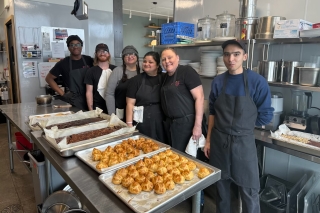 Six people in a kitchen with large sheet pans of baked goods