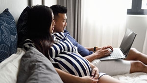 A man and pregnant woman sitting on a bed looking at their laptop