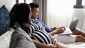 Social Media Toolkit: Share How Clients Can Cover More of Their Salaries A man and pregnant woman sitting on a bed looking at their laptop