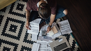 Thumbnail of overhead image of man sitting on floor with laptop surrounded by bills.
